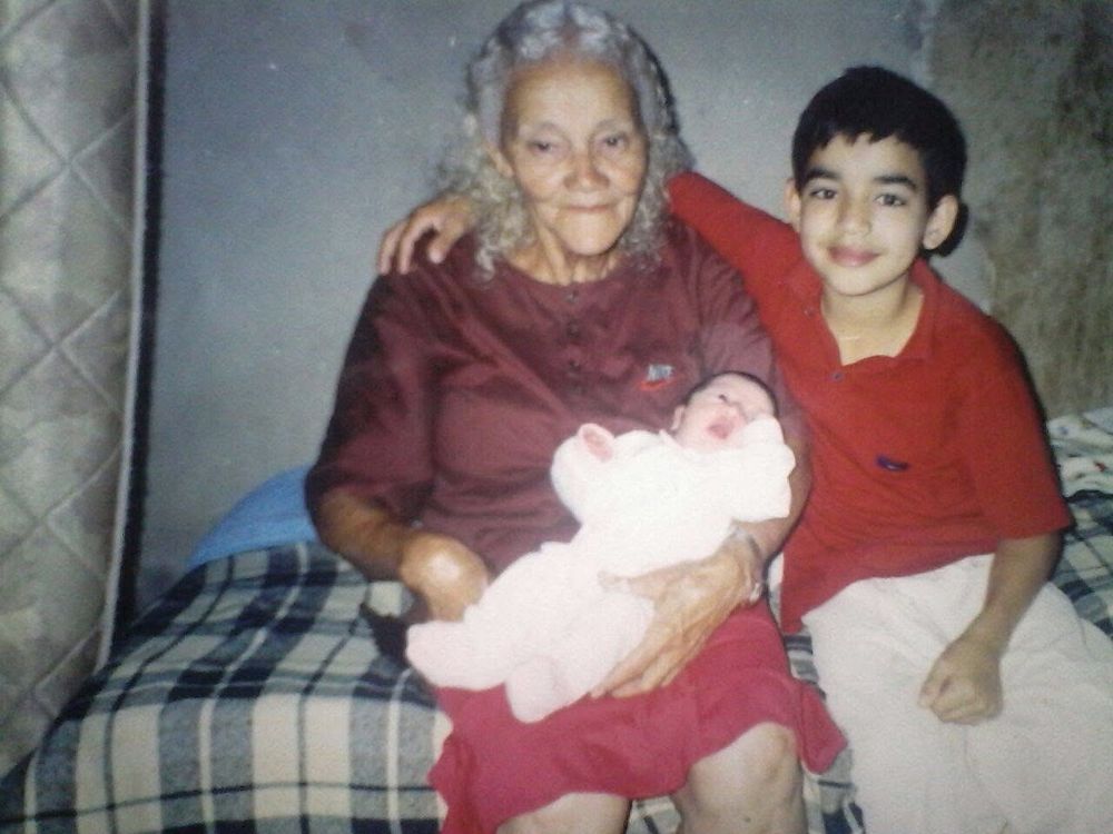 Fotografia antiga de um menino de camisa vermelha ao lado de uma senhora de cabelos grisalhos e cacheados, que segura um bebê envolto em manta clara, sentados sobre uma cama com cobertor xadrez