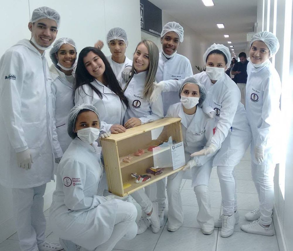 Grupo de estudantes de jaleco branco, touca e máscara posando num corredor, com uma caixa de madeira que contém modelos de peças dentárias