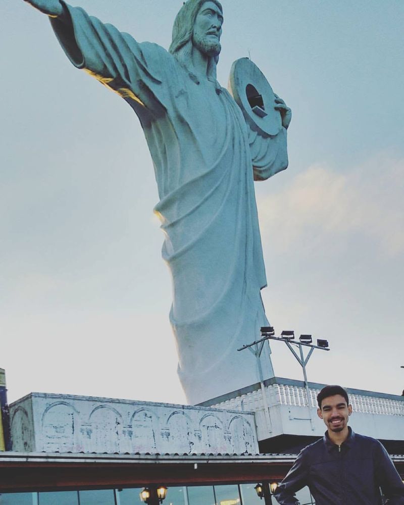 Um jovem de jaqueta escura sorri ao pé da estátua do Cristo Luz, em Balneário Camboriú, com o céu ao fundo