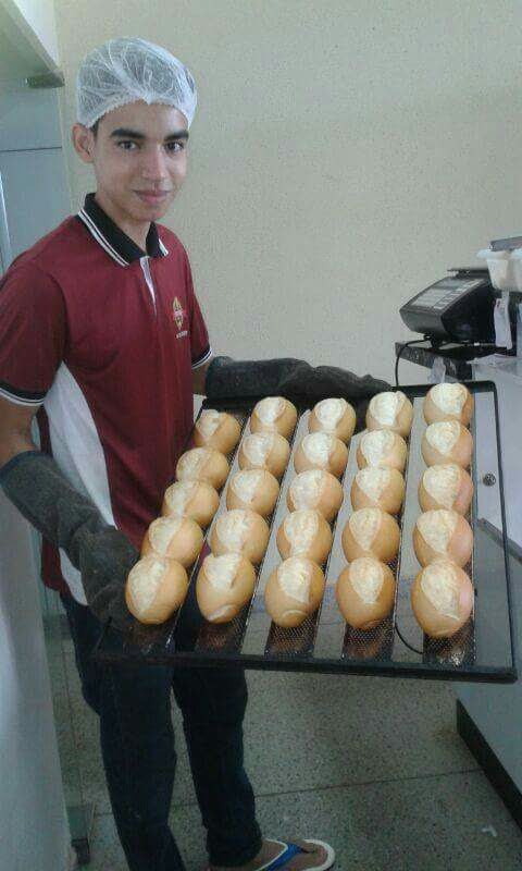 Jovem de touca descartável e camisa polo vinho, sorrindo, segura uma assadeira com pãezinhos franceses recém-assados, usando luvas térmicas, em uma cozinha de panificadora